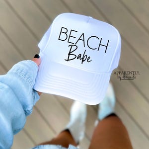 May include: White trucker hat with black text that reads "Beach Babe". The hat is held by a person wearing a light blue denim shirt.