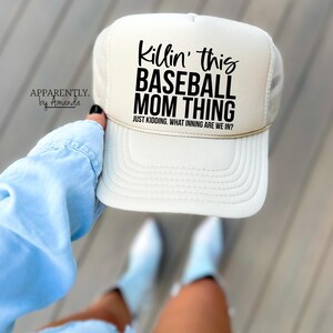 Op de afbeelding: Cr&egrave;mekleurige baseballcap met een mesh achterkant en de tekst "Killin' this BASEBALL MOM THING" in het zwart. Daaronder staat "JUST KIDDING. WHAT INNING ARE WE IN?". De pet wordt door een persoon omhooggehouden.