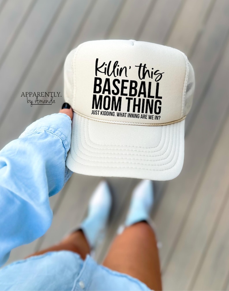 Op de afbeelding: Cr&egrave;mekleurige baseballcap met een mesh achterkant en de tekst "Killin' this BASEBALL MOM THING" in het zwart. Daaronder staat "JUST KIDDING. WHAT INNING ARE WE IN?". De pet wordt door een persoon omhooggehouden.