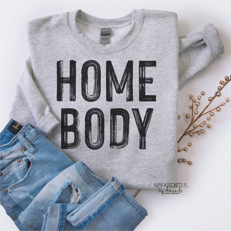 May include: Gray sweatshirt with the text "HOME BODY" in black brushstroke lettering. The text "APPARENTLY. by Amanda" is printed in black at the bottom of the sweatshirt.