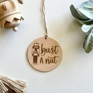 May include: A round wooden ornament with a laser-etched nutcracker design and the phrase "bust a nut." The ornament hangs from a natural twine string. The background includes a wooden nutcracker and greenery.