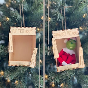 May include: Two wooden Christmas tree ornaments. One is empty and labeled "ELF TREE HOUSE". The other ornament is labeled "SNOWFLAKE'S TREE HOUSE" and contains a green Grinch figure wearing a red suit and hat. Both ornaments are hanging on a Christmas tree.