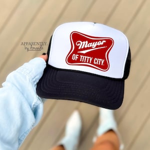 May include: A black and white trucker hat with a red and white graphic that reads "Mayor of Titty City." The hat has a white front panel, a black brim, and a black mesh back. The text "Apparently by Amanda" is printed on the side.