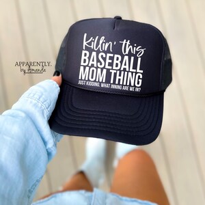 Op de afbeelding: Een zwarte baseballcap met witte tekst met de tekst "Killin' this BASEBALL MOM THING" en "JUST KIDDING. WHAT INNING ARE WE IN?". De cap heeft een mesh achterkant en een gebogen klep. De merknaam "APPARENTLY. by Amanda" is ook zichtbaar.