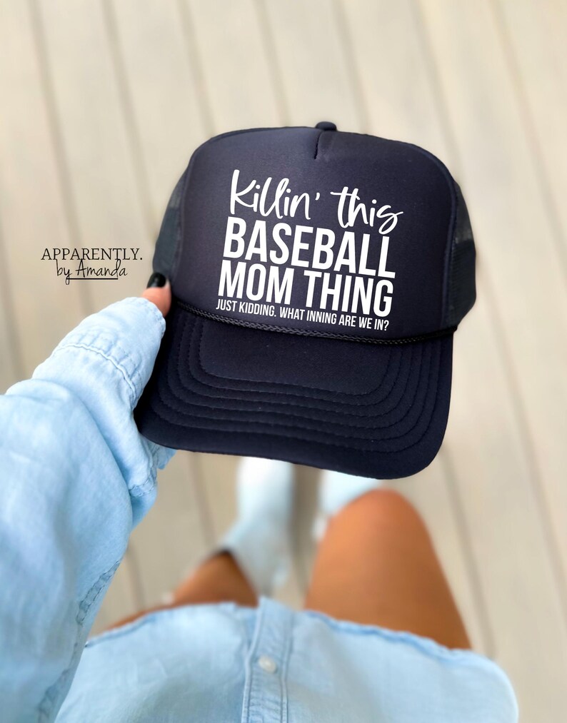 Op de afbeelding: Een zwarte baseballcap met witte tekst met de tekst "Killin' this BASEBALL MOM THING" en "JUST KIDDING. WHAT INNING ARE WE IN?". De cap heeft een mesh achterkant en een gebogen klep. De merknaam "APPARENTLY. by Amanda" is ook zichtbaar.
