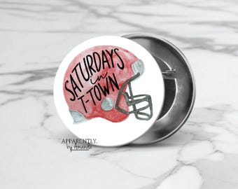 May include: A white button with a red and white football helmet and the text "Saturdays in T-Town" in black. The button also has the text "Apparently. by Amanda" in black.