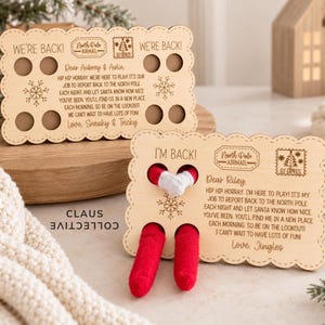 May include: Two rectangular light-colored wooden signs with scalloped edges. The top sign reads WE'RE BACK! and features North Pole Airmail branding. The bottom sign reads I'M BACK! and has two holes with red fabric elf legs and white mittens protruding.