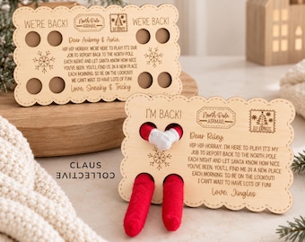 May include: Two rectangular light-colored wooden signs with scalloped edges. The top sign reads WE'RE BACK! and features North Pole Airmail branding. The bottom sign reads I'M BACK! and has two holes with red fabric elf legs and white mittens protruding.