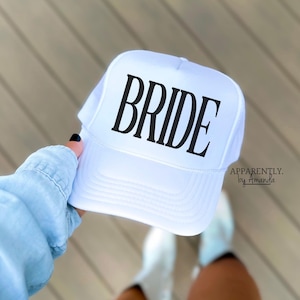 May include: White trucker hat with black text that reads "BRIDE" on the front. The hat is being held by a person wearing a light blue shirt.