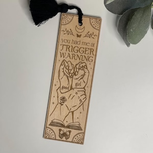 May include: Wooden bookmark with a black tassel. The bookmark features a hand-drawn illustration of two hands clasped together with the text "you had me at trigger warning" and the words "good girl" written on the hands. The bookmark also features a butterfly and a crescent moon.