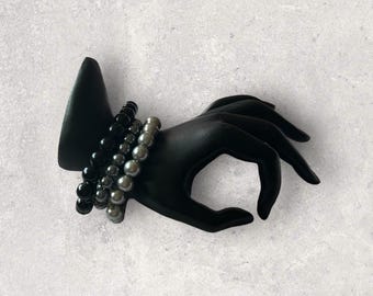 May include: A black hand-shaped jewellery display holding three beaded bracelets. The bracelets feature black and grey beads. The display is on a light grey surface.