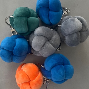 May include: A set of seven colourful, plush, knotted ball keyrings. The keyrings are in shades of blue, green, grey, and orange.