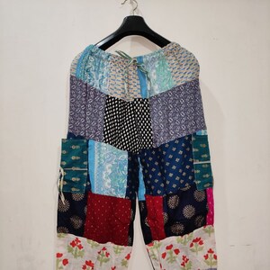 Pode incluir: Calças de patchwork com uma variedade de quadrados de tecido em azul, vermelho, branco e preto com diversos padrões florais, geométricos e de pontos. As calças têm uma cintura elástica com cordão e punhos elásticos nos tornozelos.