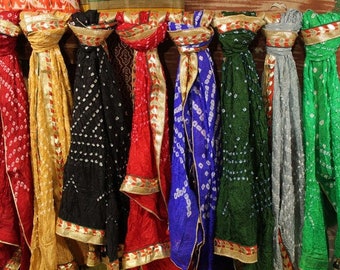 May include: A row of eight colorful scarves featuring a tie-dye bandhani pattern with small white dots. Each scarf is draped over a horizontal bar and finished with a wide, metallic gold decorative border along the edges and the top hem.