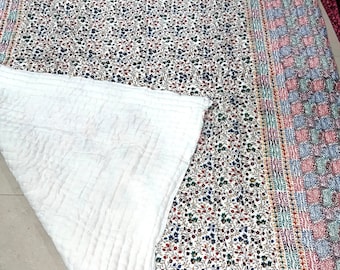 May include: A rectangular quilt featuring a white base with a dense floral pattern in red, blue, and green. The border displays a checkered pattern in muted pink, blue, and green tones. The underside is a plain white fabric with visible stitching.