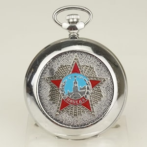 Puede incluir: Un reloj de bolsillo plateado con una esfera detallada. La esfera del reloj presenta una estrella roja con un emblema central y la palabra "Pobeda" en letras rojas. El reloj tiene una tija de cuerda plateada y una caja circular.