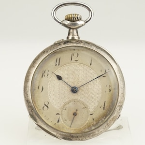 Puede incluir: Un reloj de bolsillo plateado con esfera blanca y agujas negras. El reloj tiene un pequeño dial de segundos y un centro texturizado. La parte superior tiene un lazo para una cadena y una corona dorada. El reloj es antiguo.