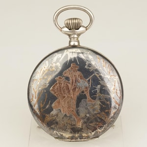 Puede incluir: Reloj de bolsillo plateado con una escena grabada de dos figuras con sombreros. La esfera del reloj tiene un fondo oscuro con detalles dorados y cobrizos. El reloj tiene una tija de cuerda plateada y una parte superior decorativa.