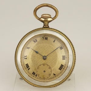 Puede incluir: Un reloj de bolsillo dorado con una esfera dorada texturizada, números romanos negros y manecillas negras. El reloj tiene un borde blanco de nácar y un lazo dorado en la parte superior. Un reloj clásico.