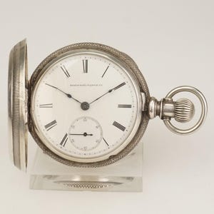 Könnte beinhalten: Silberne Taschenuhr mit weißem Zifferblatt und schwarzen römischen Ziffern. Die Uhr hat ein kleines Sekunden-Zifferblatt und die Worte "ELGIN RAIL WATCH CO" sind auf dem Zifferblatt aufgedruckt. Die Uhr ist geöffnet.