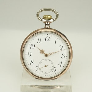 Puede incluir: Un reloj de bolsillo plateado y dorado con esfera blanca y números negros. El reloj tiene un pequeño dial de segundos y un lazo plateado para una cadena. El reloj se exhibe sobre un soporte transparente.