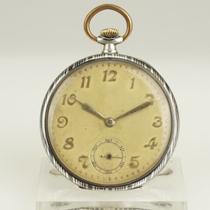 Puede incluir: Un reloj de bolsillo vintage con caja plateada y esfera crema. El reloj presenta números negros, manecillas y un pequeño dial de segundos. Tiene un vástago de cuerda dorado y un lazo para una cadena.