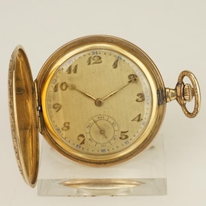 Puede incluir: Un reloj de bolsillo dorado con la tapa abierta, que revela una esfera crema con números y manecillas negras. El reloj tiene un pequeño dial de segundos y una tija de cuerda. El reloj se exhibe sobre un soporte transparente.