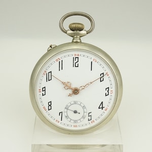 Puede incluir: Un reloj de bolsillo plateado con esfera blanca y números negros. El reloj tiene un pequeño dial de segundos y agujas doradas. El reloj mide aproximadamente 5 cm de diámetro.