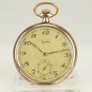 Puede incluir: Un reloj de bolsillo vintage con esfera crema, números y agujas negros. El reloj tiene un bisel dorado, un pequeño dial de segundos y una corona dorada. La marca "Zentra" está impresa en la esfera.