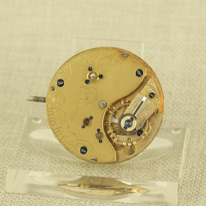 Puede incluir: Primer plano de un movimiento de reloj antiguo. El mecanismo dorado presenta intrincados engranajes, tornillos y un volante con una joya azul. La esfera tiene una escritura grabada y el número 83208. Un pequeño pasador metálico se extiende desde el lateral.