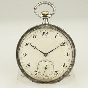 Könnte beinhalten: Eine silberne Taschenuhr mit weißem Zifferblatt und schwarzen Ziffern. Die Uhr hat ein kleines Sekunden-Zifferblatt und eine silberne Kettenschlaufe. Die Zeiger sind dunkelblau. Das Uhrengehäuse hat ein dekoratives, geätztes Design.