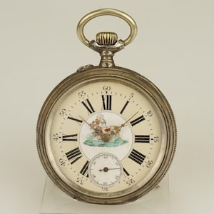 Puede incluir: Reloj de bolsillo plateado con esfera crema y números romanos negros. El reloj presenta una pequeña subesfera y una escena pintada de una persona en un carro tirado por un perro. Tiene una corona de cuerda y un asa plateadas.