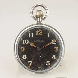 Puede incluir: Un reloj de bolsillo vintage con esfera negra y caja plateada. El reloj presenta números y agujas doradas, con un pequeño dial de segundos en la parte inferior. La marca "Jaeger-LeCoultre" es visible. Un lazo plateado en la parte superior.