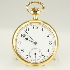 Puede incluir: Un reloj de bolsillo dorado con esfera blanca y números negros. El reloj tiene un pequeño dial de segundos y un lazo dorado en la parte superior. Las manecillas son negras y el reloj está sobre un fondo blanco.