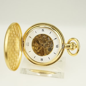 Puede incluir: Un reloj de bolsillo dorado con esfera abierta, que revela los intrincados mecanismos internos. El reloj tiene una esfera blanca con números romanos y manecillas negras. La tapa abierta muestra un patrón texturizado. El reloj está sobre un soporte transparente.