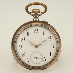 Puede incluir: Un reloj de bolsillo vintage con esfera blanca, números negros y manecillas doradas. El reloj tiene una caja plateada y dorada y un pequeño dial de segundos. La parte superior presenta una corona dorada y un lazo plateado.