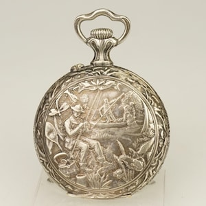 Puede incluir: Reloj de bolsillo plateado con una escena en relieve detallada de un pescador. El reloj presenta un borde floral decorativo y un lazo para una cadena. La caja del reloj tiene aproximadamente 5 cm de diámetro.