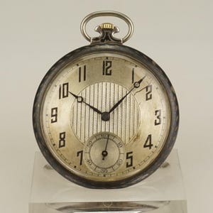Puede incluir: Un reloj de bolsillo plateado con esfera redonda y números negros. El reloj tiene un pequeño dial de segundos en la parte inferior y un centro texturizado. El reloj tiene un lazo de cadena plateado en la parte superior.