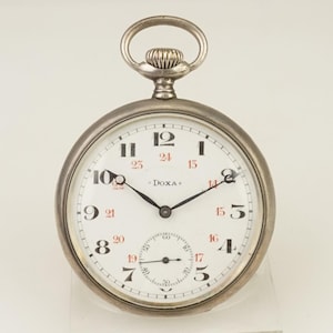 Puede incluir: Un reloj de bolsillo plateado con esfera blanca, manecillas negras y números rojos. El reloj tiene un pequeño dial de segundos y la marca "DOXA" está impresa en la esfera. El reloj tiene un anillo en la parte superior para sujetar una cadena.