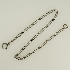 Puede incluir: Una cadena plateada con un patrón alterno de eslabones ovalados y circulares. La cadena tiene un cierre en cada extremo, uno es un anillo simple y el otro un anillo de resorte. La cadena se muestra sobre un fondo blanco.