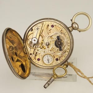 Puede incluir: Un reloj de bolsillo antiguo abierto, que revela los intrincados mecanismos internos. El reloj tiene una caja plateada y un interior dorado con engranajes, joyas y una pequeña esfera secundaria. Una llave está adjunta.