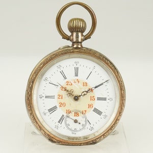 Puede incluir: Un reloj de bolsillo antiguo con esfera blanca, números romanos negros y números arábigos rojos. El reloj tiene un bisel dorado con un diseño ornamentado y una caja plateada. Incluye un pequeño dial de segundos y una corona.