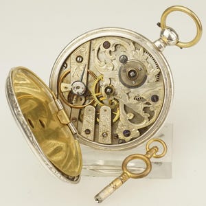 Puede incluir: Un reloj de bolsillo plateado abierto con una llave dorada. Los intrincados mecanismos internos del reloj son visibles, incluyendo engranajes y grabados decorativos. La caja del reloj está abierta, revelando los detalles. La llave descansa junto al reloj.