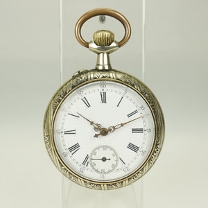 Könnte beinhalten: Eine antike Taschenuhr mit weißem Zifferblatt, schwarzen römischen Ziffern und filigranen Zeigern. Das silberfarbene Gehäuse weist aufwendige Gravurmuster und eine goldfarbene Krone und einen Bügel auf. Ein kleineres Hilfszifferblatt befindet sich unten auf dem Zifferblatt.