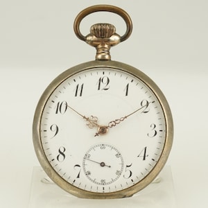 Puede incluir: Un reloj de bolsillo plateado con esfera blanca y números negros. El reloj tiene delicadas manecillas doradas y un pequeño dial de segundos. La parte superior presenta un lazo y una corona decorativa. El reloj mide aproximadamente 5 cm de diámetro.