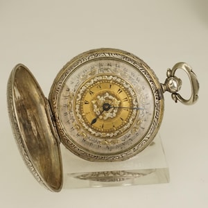 Puede incluir: Un reloj de bolsillo antiguo con la tapa abierta. El reloj presenta una caja plateada con intrincados grabados y una esfera dorada con números arábigos. Las manecillas del reloj son azules. El reloj se exhibe sobre un soporte transparente.