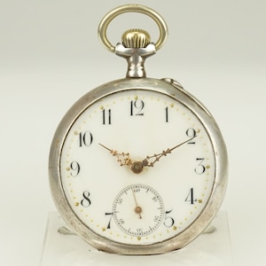 Puede incluir: Un reloj de bolsillo plateado con esfera blanca y números negros. El reloj tiene manecillas doradas y un pequeño dial de segundos. La caja es plateada y la corona dorada. Un reloj clásico.