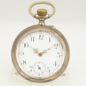 Puede incluir: Un reloj de bolsillo vintage con esfera blanca, números negros y delicadas manecillas doradas. La caja es plateada y tiene un pequeño dial de segundos. La corona es dorada y tiene un asa para una cadena. Diámetro: aproximadamente 5 cm.