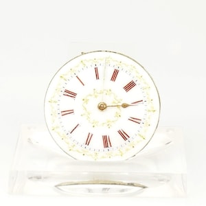 Puede incluir: Una esfera de reloj blanca con números romanos rojos y manecillas doradas. La esfera tiene un diseño floral dorado decorativo en el borde. El reloj se muestra sobre un soporte transparente y rectangular.