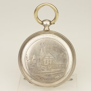 Puede incluir: Un reloj de bolsillo plateado con un anillo dorado. La esfera del reloj presenta una escena grabada de un edificio con dos agujas y la palabra "ALTENBRUCH" en la parte inferior. El reloj tiene forma circular.
